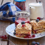 A layered puff pastry cake with vanilla cream and jam in between, called as Mille-Feuille pastry on wooden surface, surrounded by a teapot, cherries, blueberries a towel and jam jar - an afternoon tea scene