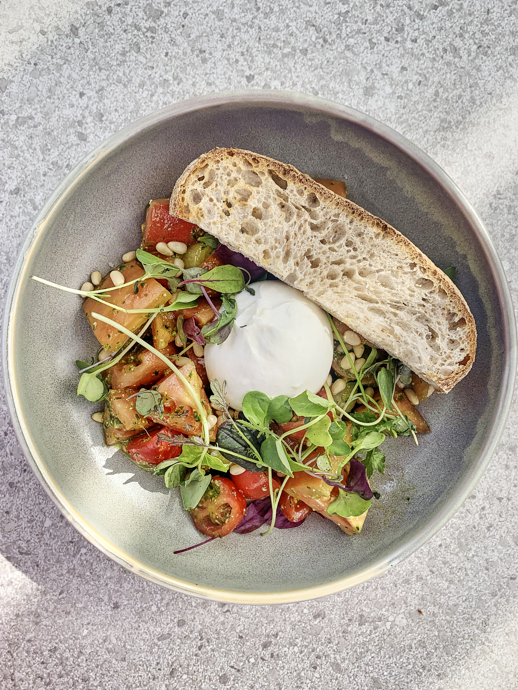 Tomato salad with burrata and a slice of crusty bread in a gray bowl on a gray patterned stone table. A sunny afternoon scene with some shadows.