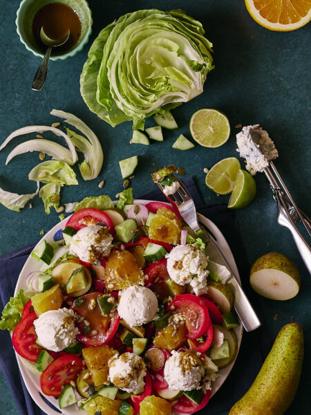 Fresh salad with mixed greens, tomatoes, cucumbers, oranges, pears, and feta cheese balls, drizzled with a dark vinaigrette, arranged on a white plate., placed on a dark blue-green backdrop