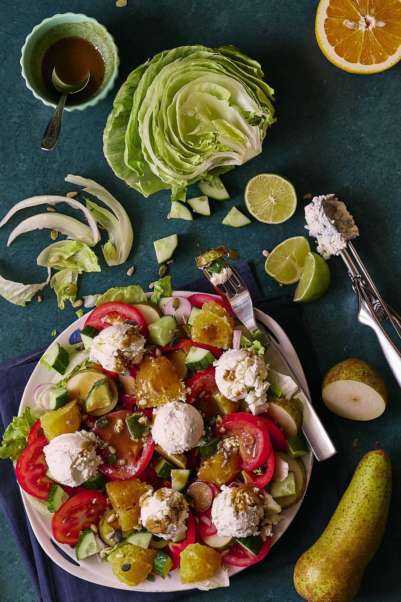 Fresh salad with mixed greens, tomatoes, cucumbers, oranges, pears, and feta cheese balls, drizzled with a dark vinaigrette, arranged on a white plate., placed on a dark blue-green backdrop