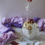 A woman's hand with red nails sprinkling shredded coconut over an oatmeal mousse dessert dish topped with pine nuts and red jam. Purple towels in the background and a wooden knife with the jam in the foreground. A candle and a candle holder in the shape of angel's wings.