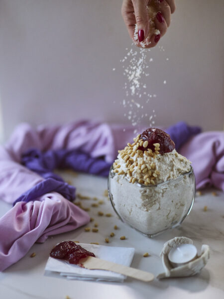 A woman's hand with red nails sprinkling shredded coconut over an oatmeal mousse dessert dish topped with pine nuts and red jam. Purple towels in the background and a wooden knife with the jam in the foreground. A candle and a candle holder in the shape of angel's wings.